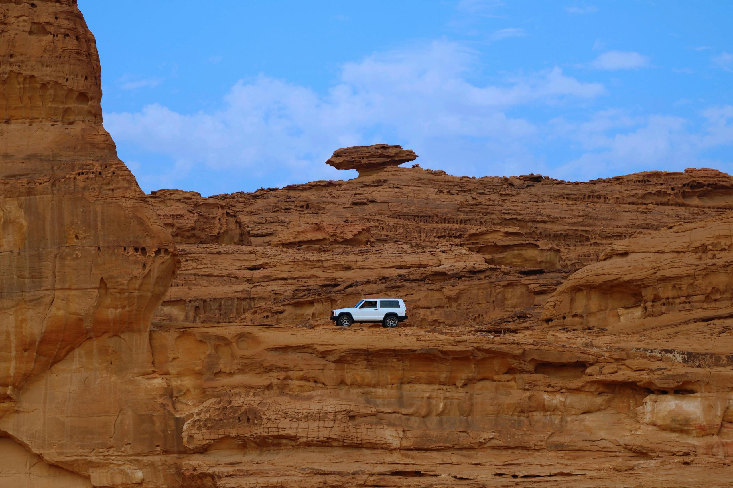 White SUV navigating sandstone cliffs in Al-Ula, showcasing rugged desert landscape.