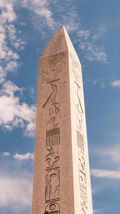 Free Tall Egyptian obelisk with hieroglyphs set against a bright blue sky and cloud backdrop. Stock Photo