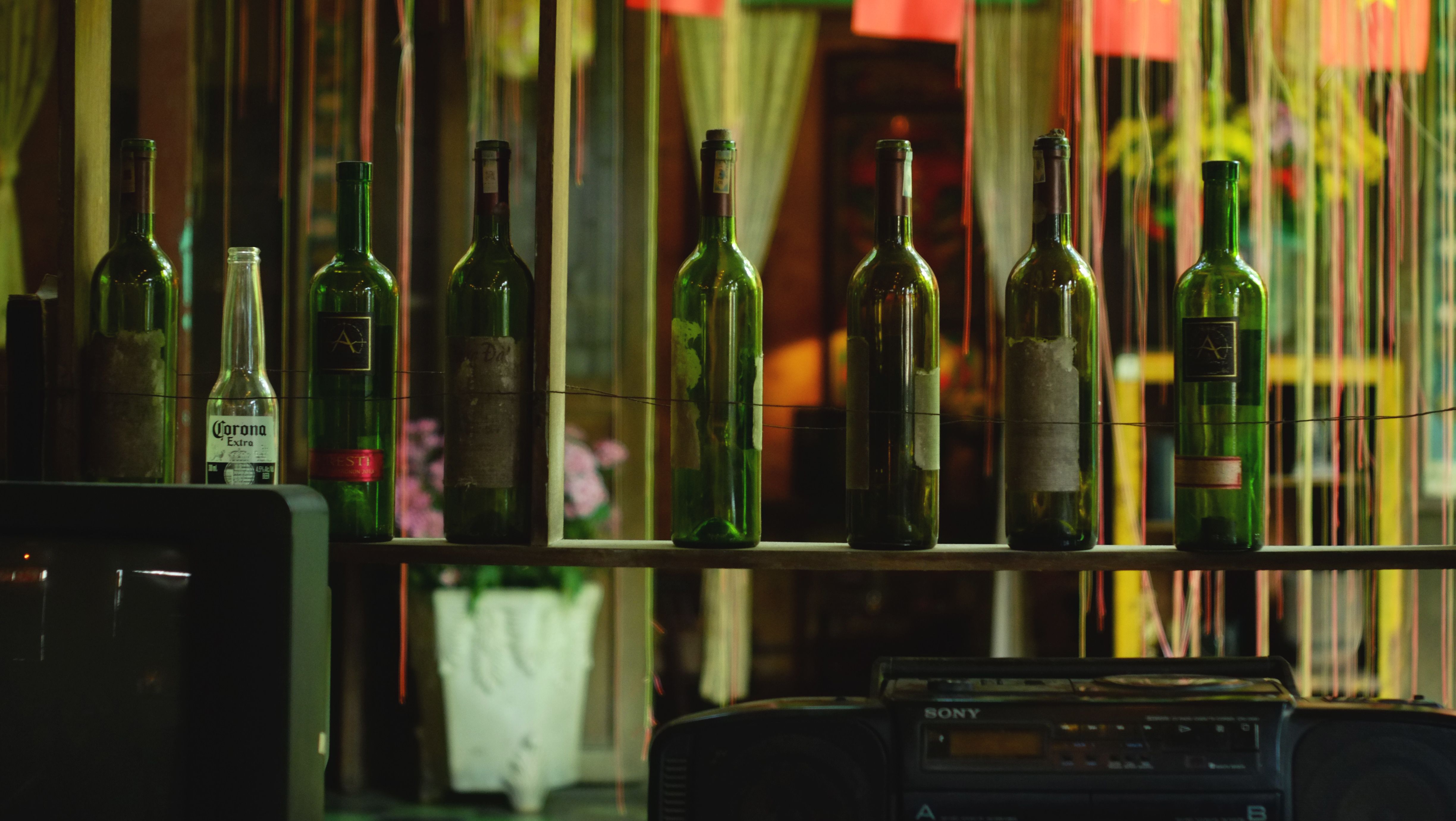 A row of green wine bottles and a Corona bottle on a wooden shelf indoors.