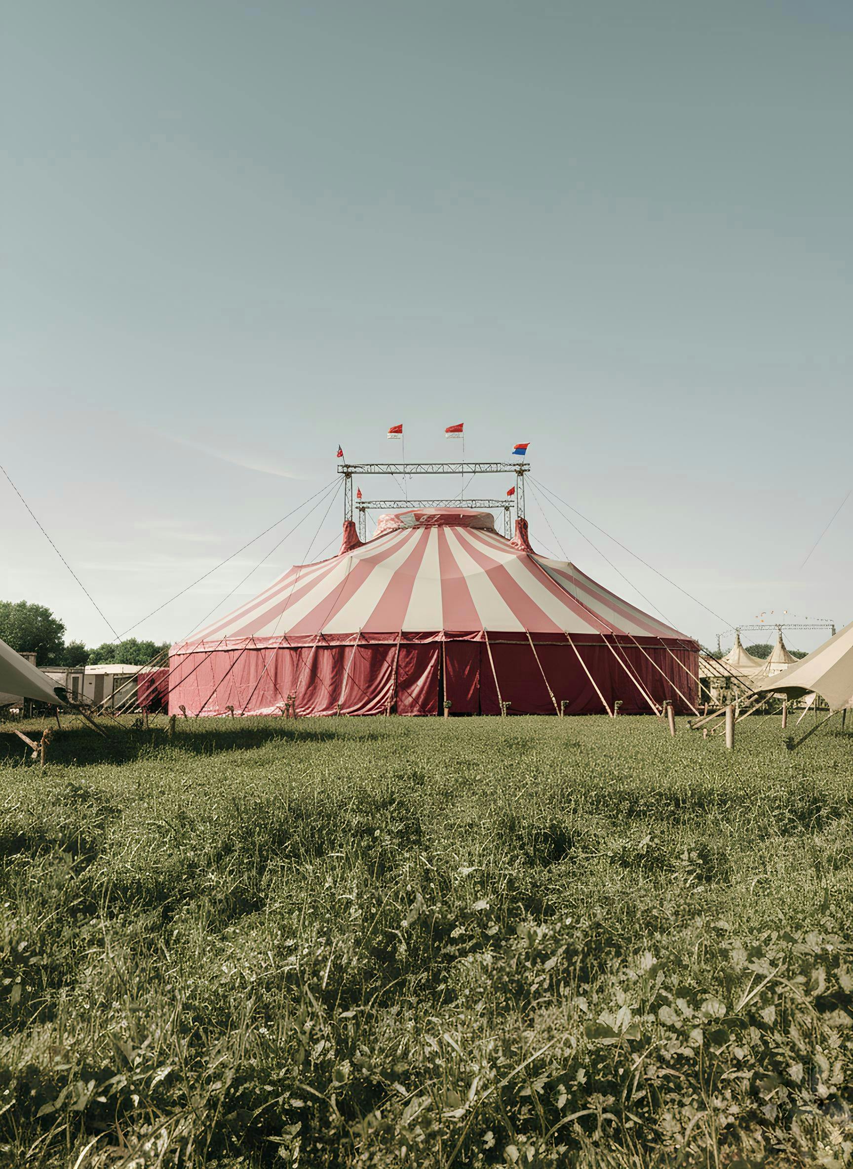A large red and white circus tent set in a green field under a clear sky.