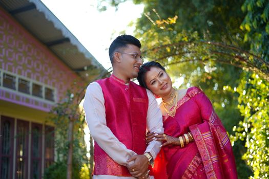 A South Asian couple in traditional attire, embracing outdoors under sunlight, smiling joyfully.