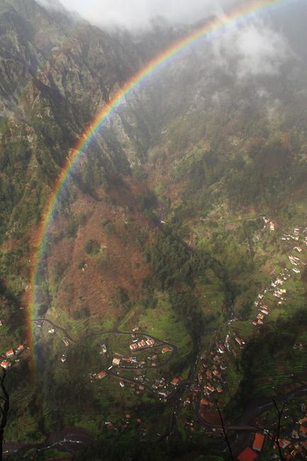 A breathtaking view of a rainbow arching over a lush mountain valley, dotted with village houses.