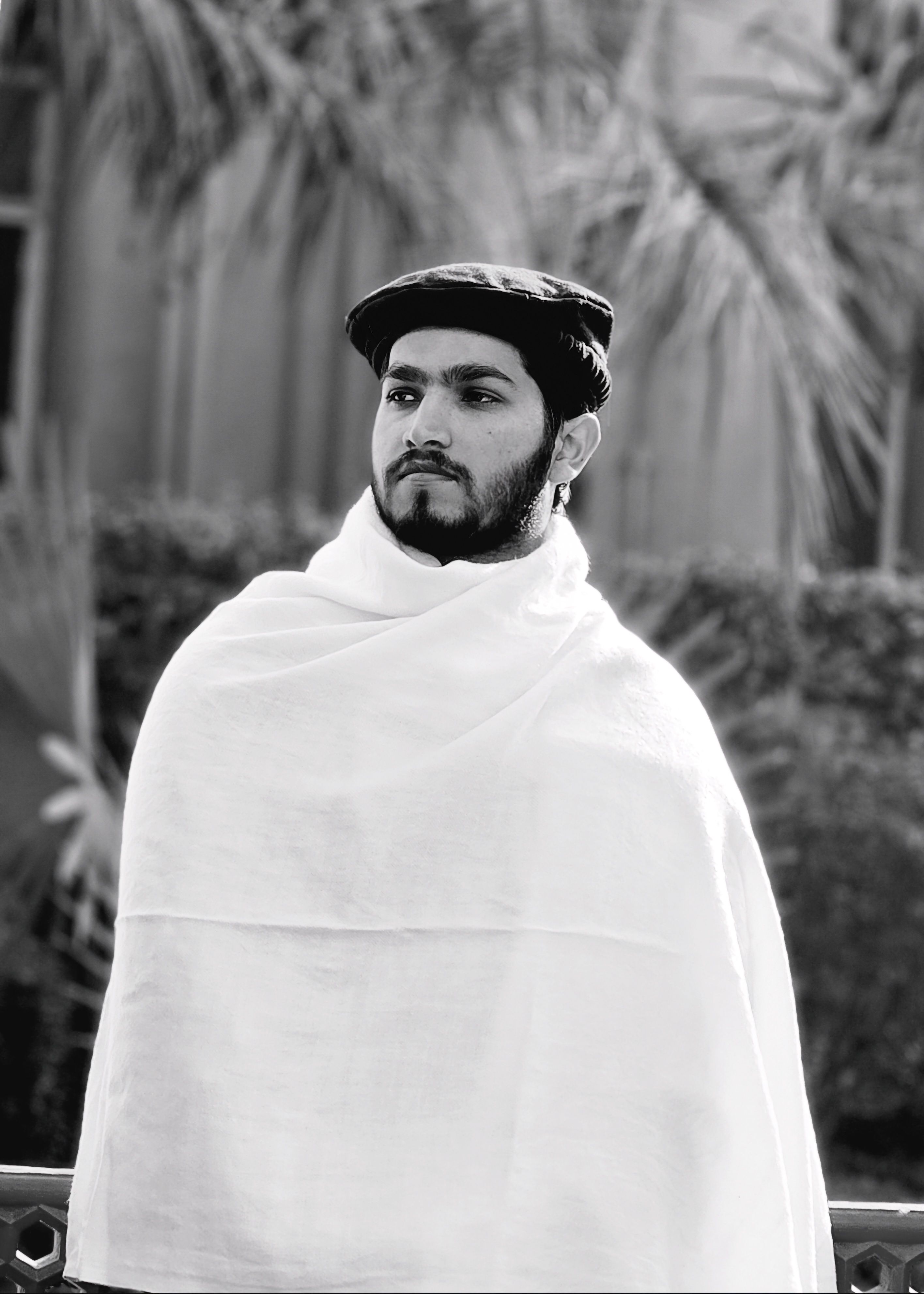 Black and white portrait of a man with a traditional cap and shawl outdoors.
