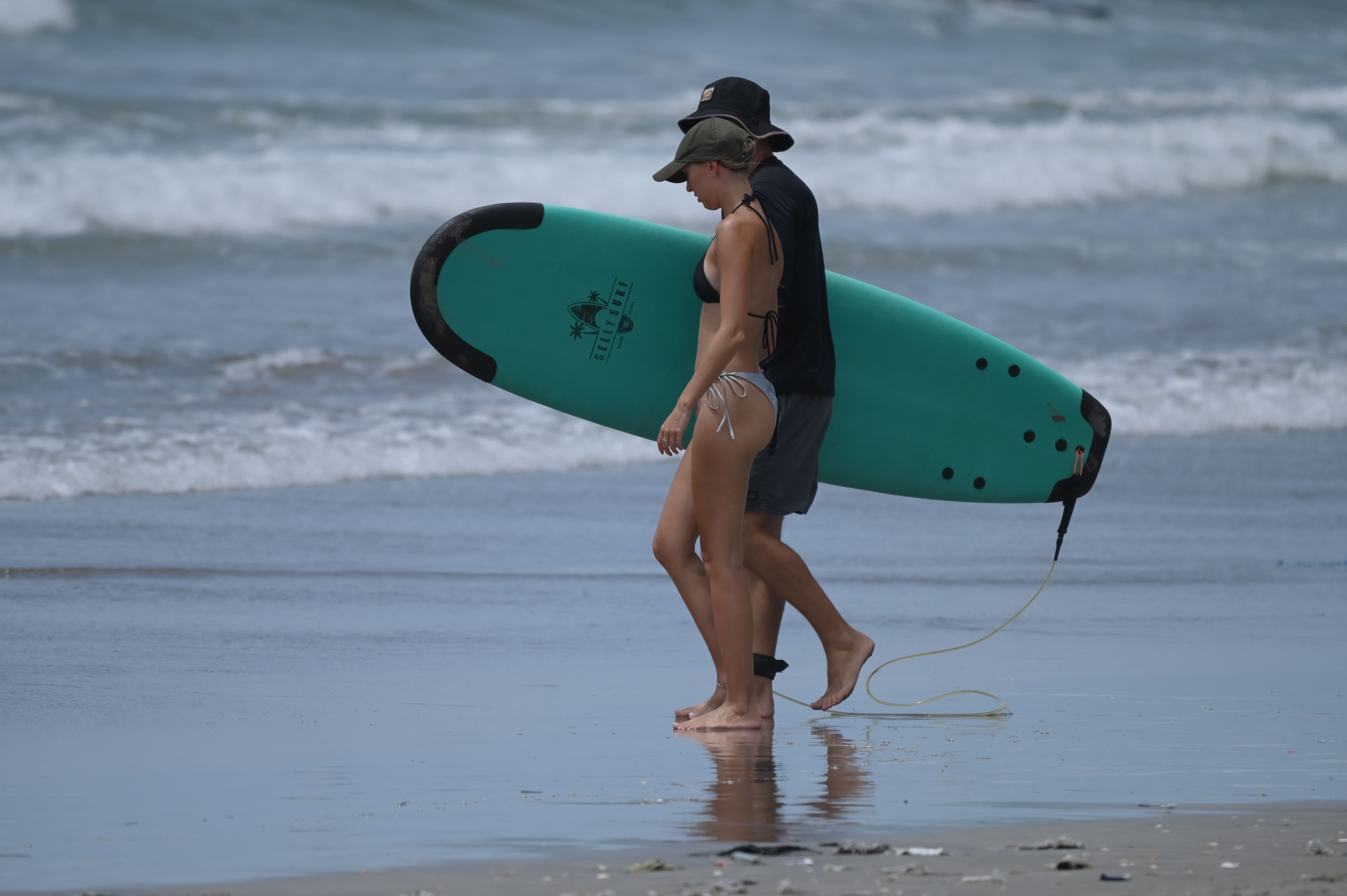 Beginner surfers carrying foam longboards on the wide grey sand of Kuta Beach in Bali