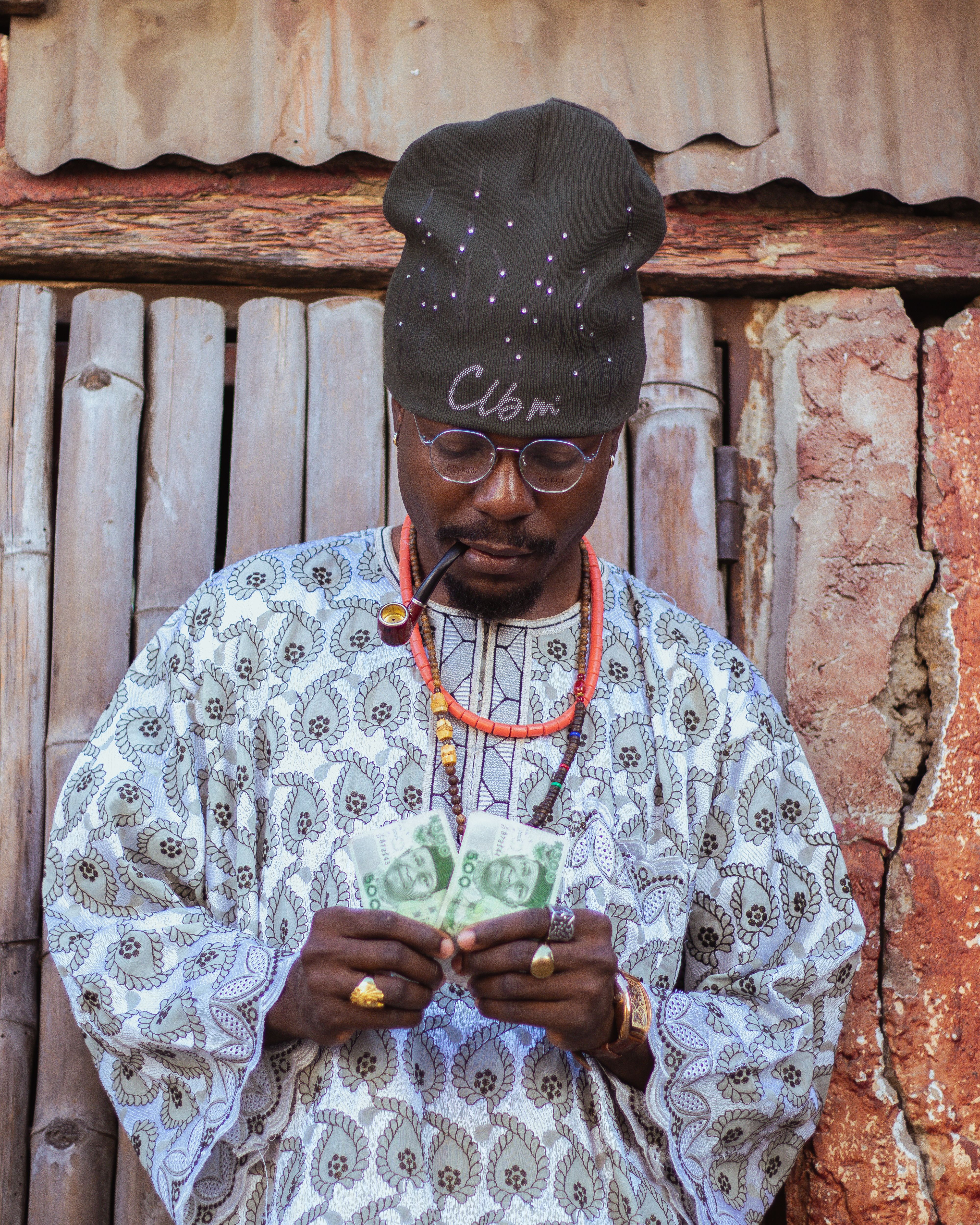 African man in traditional attire with hat and glasses counting money against wooden background.
