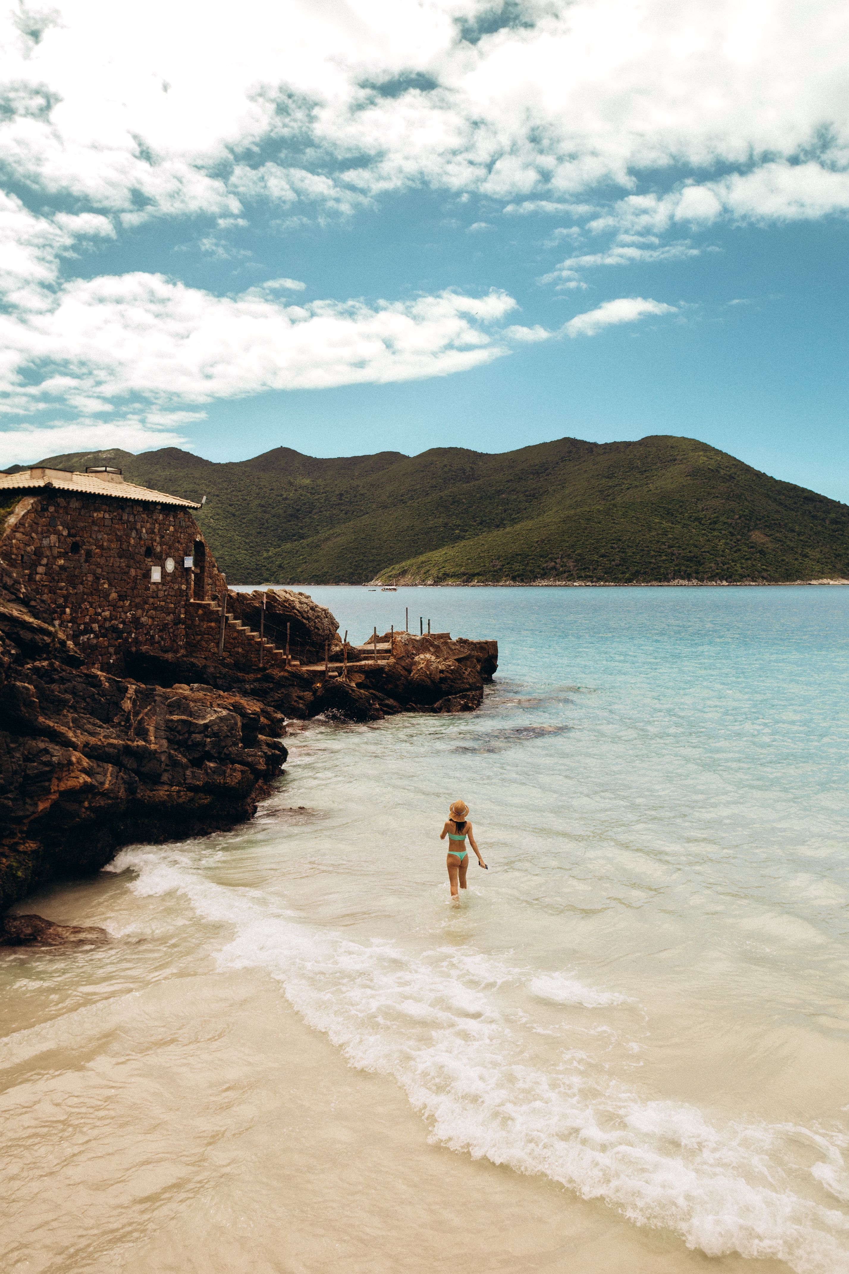 A lone child plays on a scenic beach with clear waters and lush hills under a bright sky.