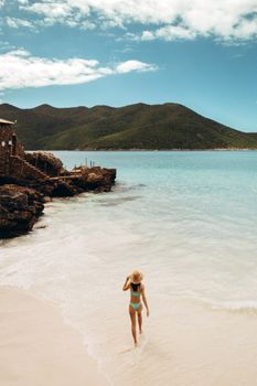 A woman in a bikini and hat walks along a serene tropical beach with lush mountains in the background.