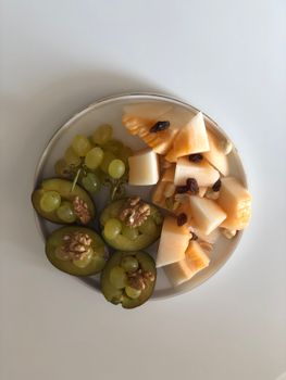 A vibrant, healthy fruit and nut platter featuring grapes, melon, plums, and walnuts on a white background.