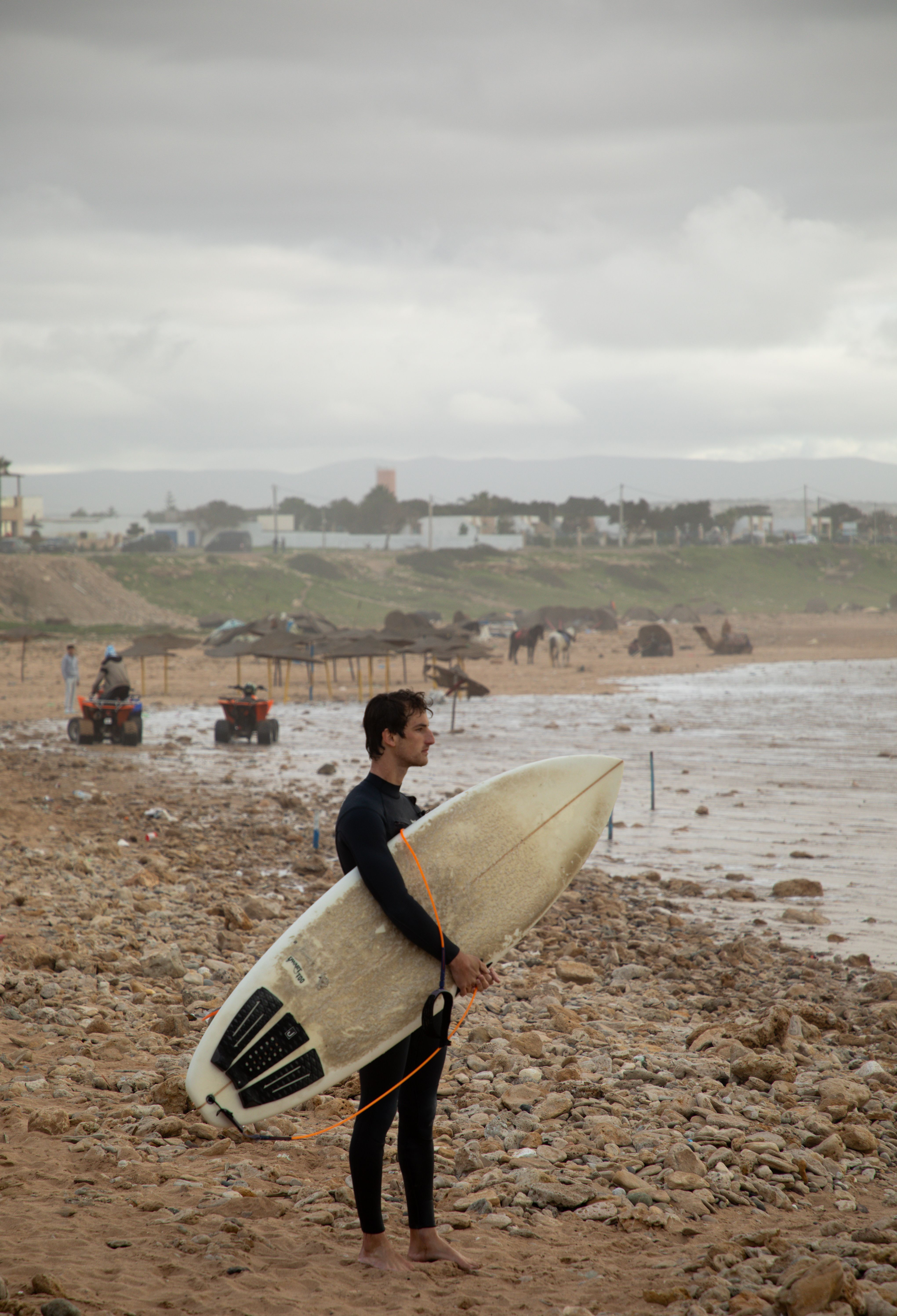 A surfer holding a surfboard stands on a rocky beach in Sidi Kaouki, Morocco, under cloudy skies.