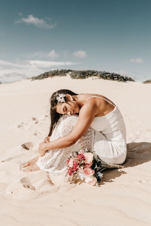 Free A woman in a white dress sits peacefully on a sandy beach with a bouquet of flowers. Stock Photo