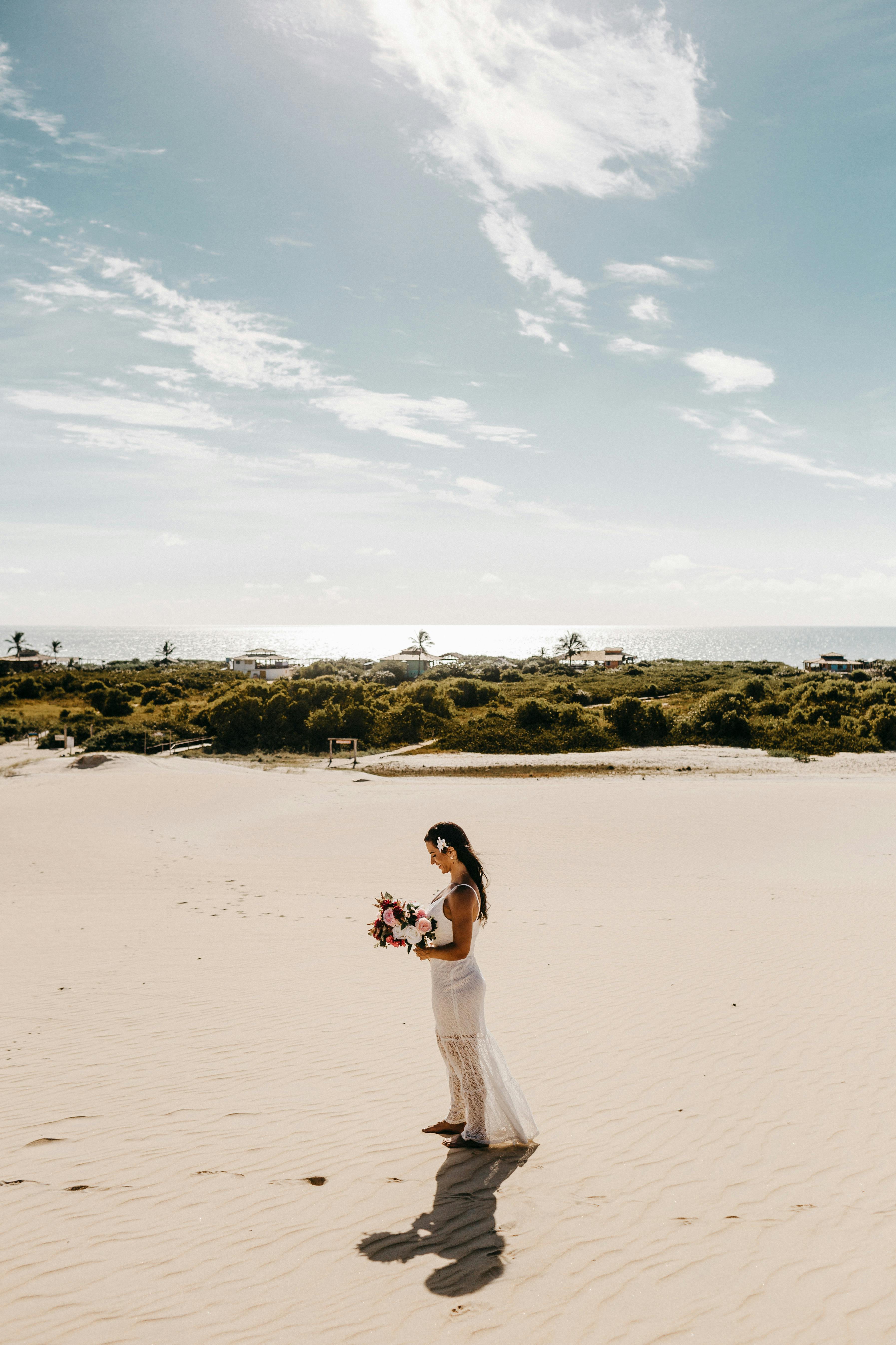 阳光明媚的海滩上，一名身穿白色连衣裙、手持鲜花的女子优雅地站着，周围环绕着自然美景