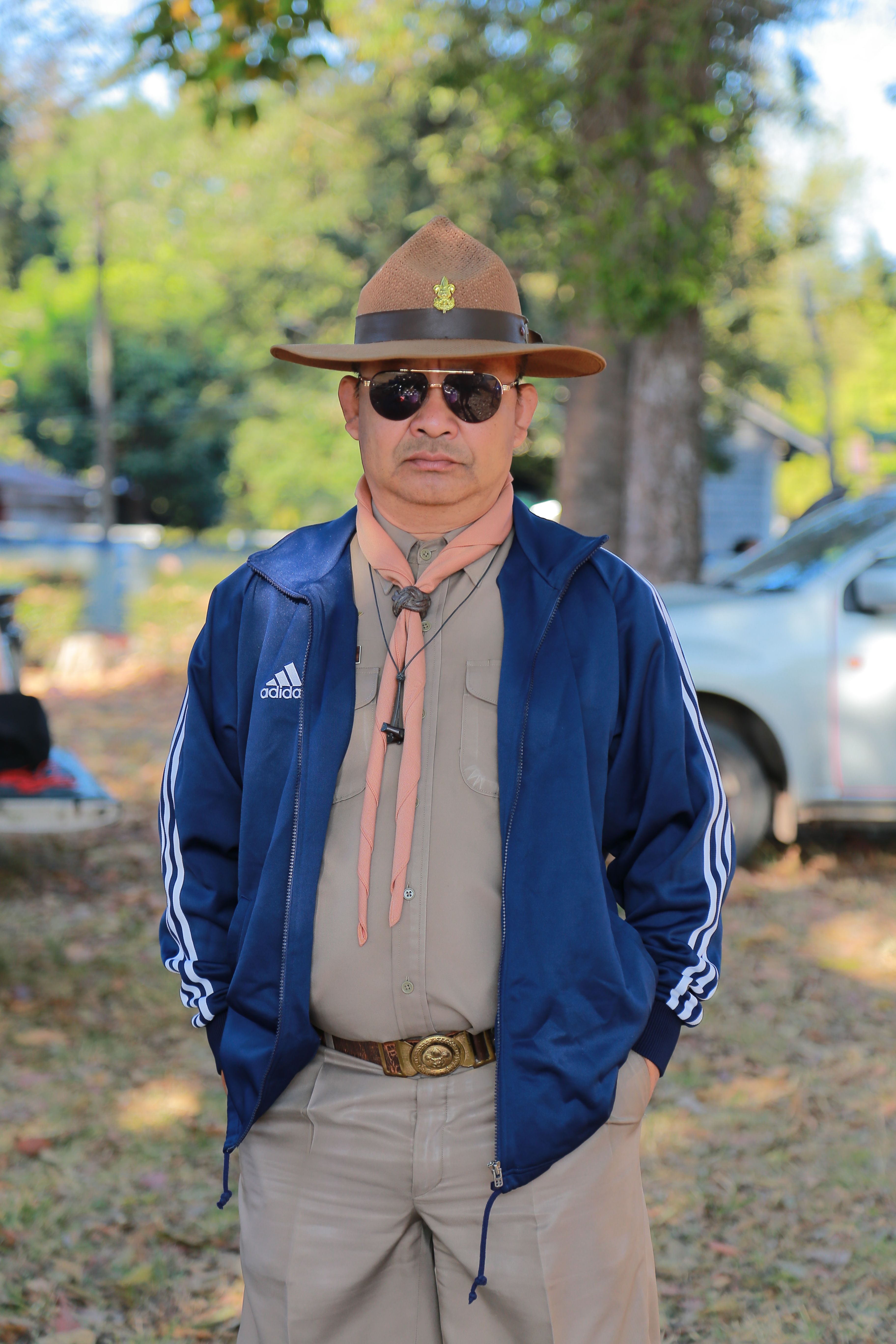 A scout leader in uniform poses outdoors, wearing sunglasses and a wide-brimmed hat.