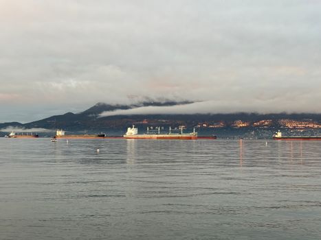 Cargo ships in Vancouver harbor with mountains and clouds at sunset, creating a serene maritime scene.
