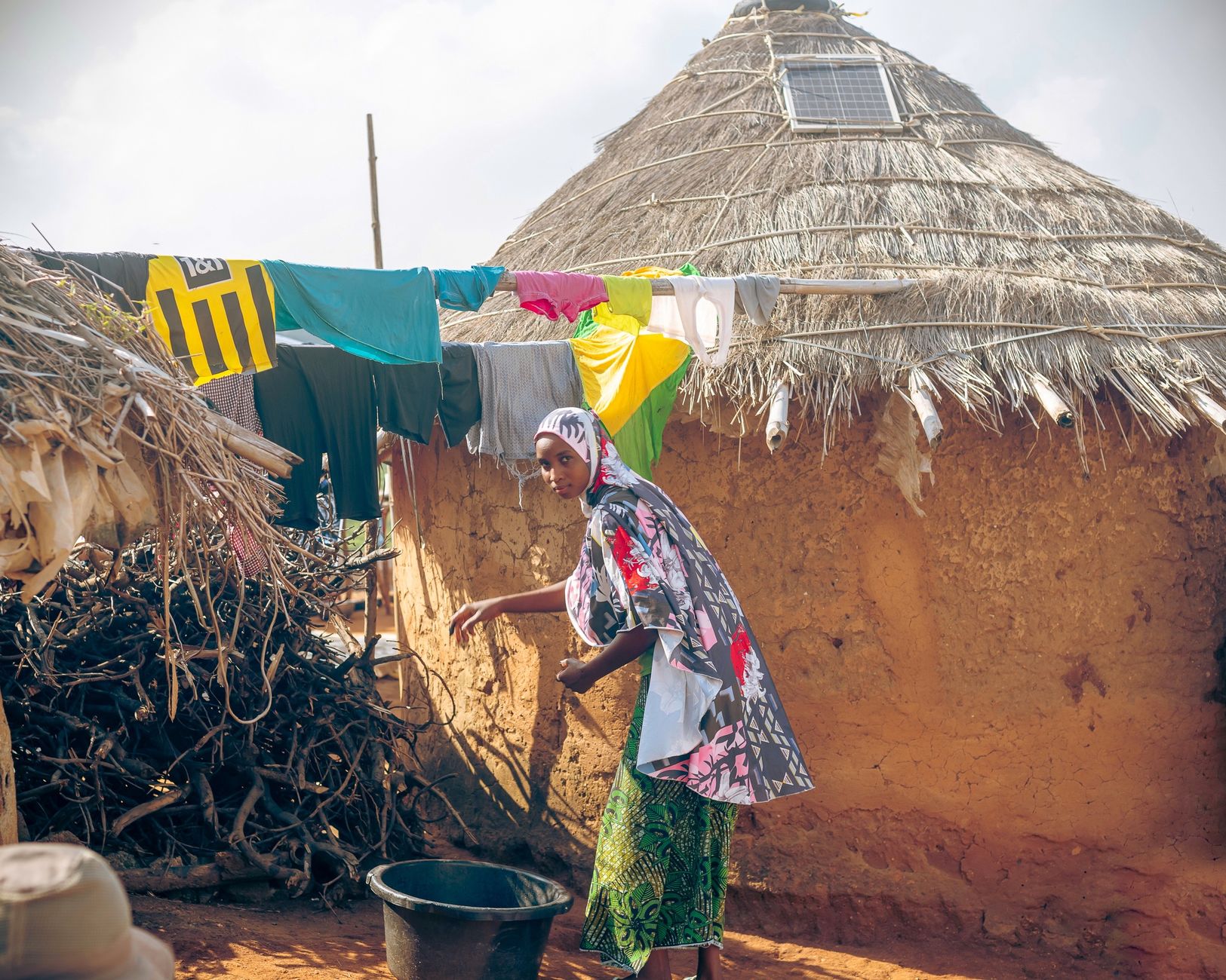 African woman in colorful attire performing daily chores outside a hut with thatched roof.