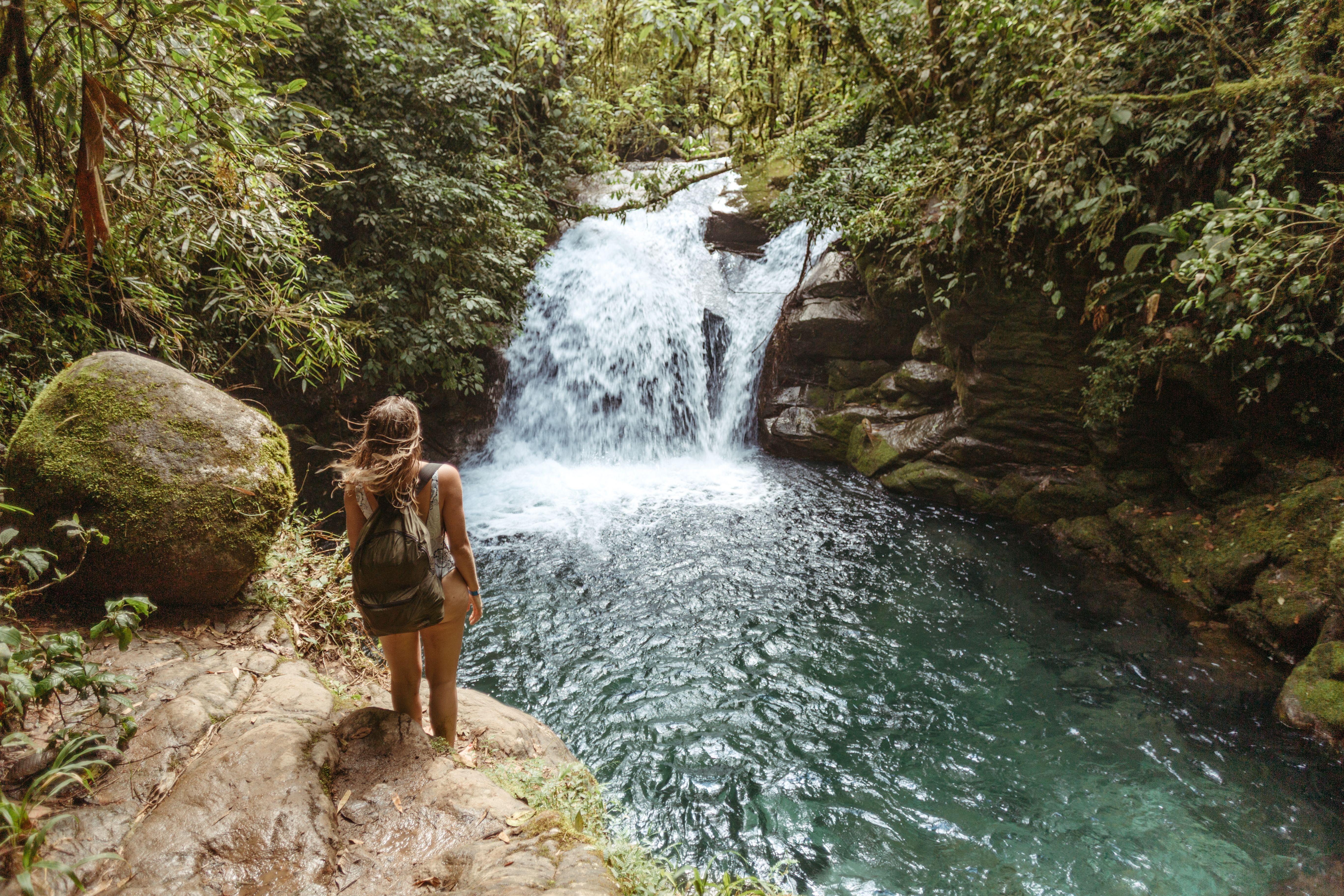 A woman with a backpack exploring a stunning tropical waterfall surrounded by lush green forest.