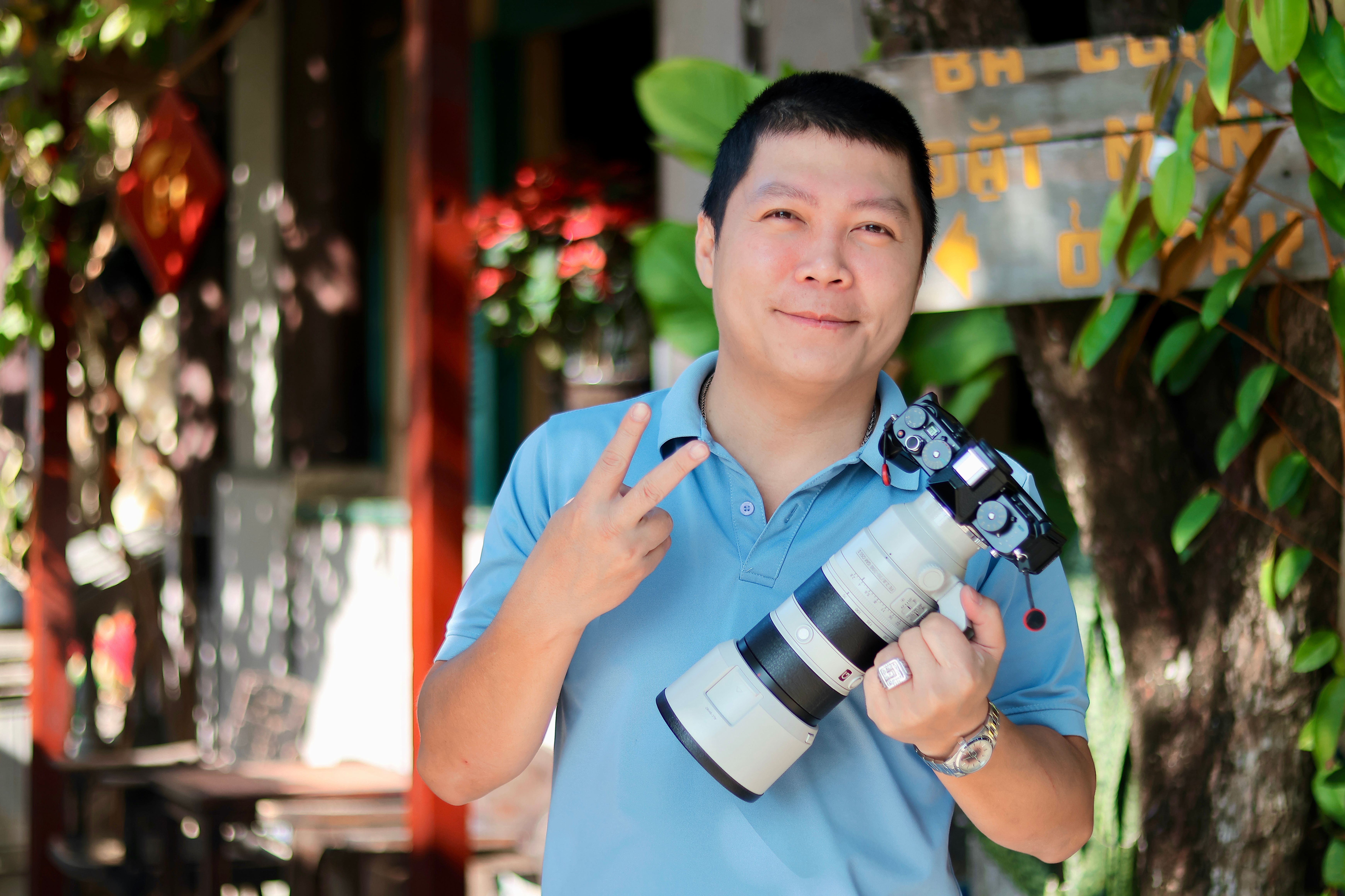 Portrait of a photographer holding a camera, making a peace sign outdoors.