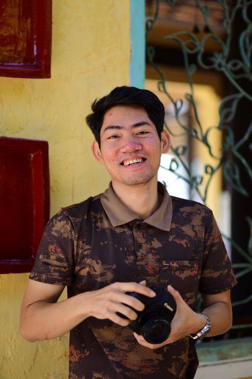 Free A cheerful man holding a camera, standing outdoors in casual attire, enjoying a sunny day. Stock Photo