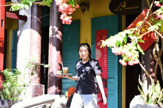 Young waitress carrying iced coffee in vibrant outdoor cafe setting with flowers.