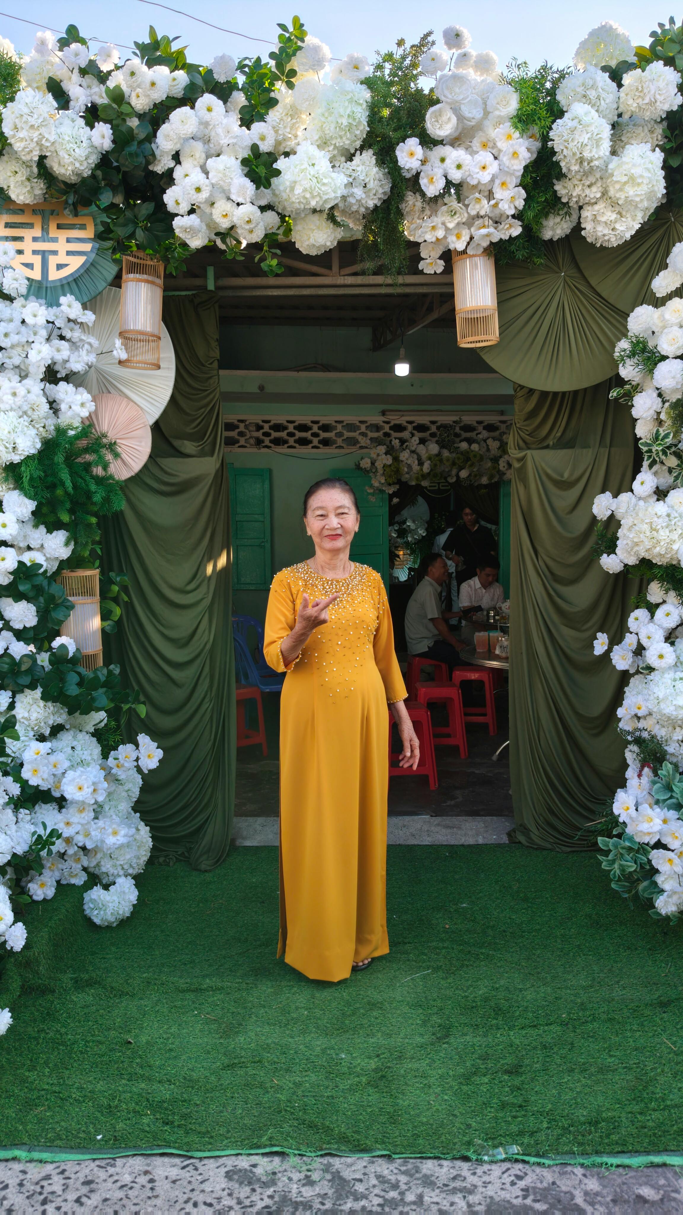 Elegant woman in yellow dress stands in floral-adorned entrance, outdoor celebration setting.