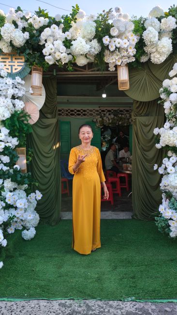 Elegant woman in yellow dress stands in floral-adorned entrance, outdoor celebration setting.