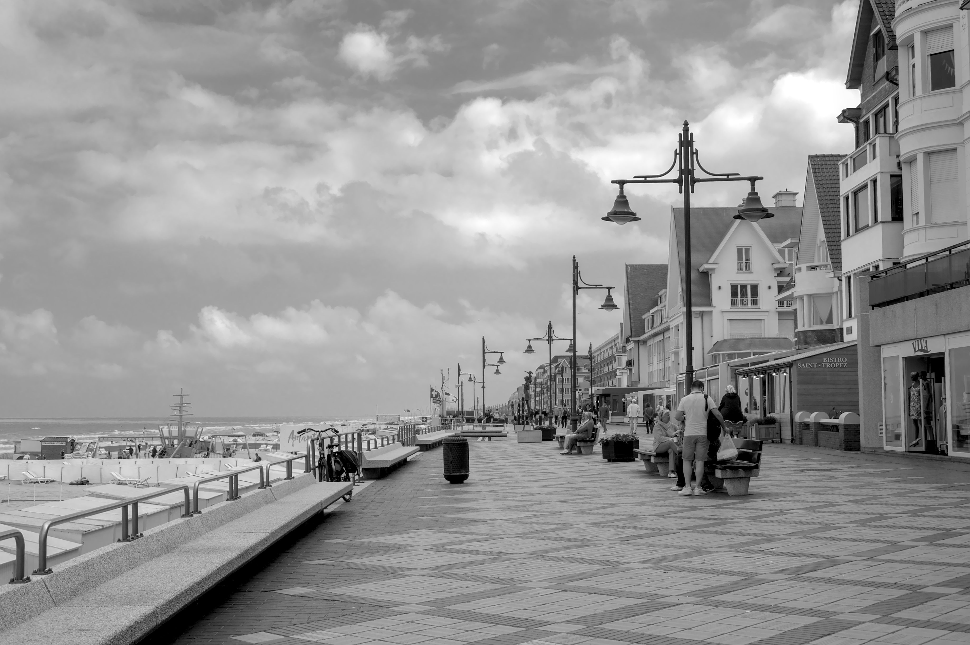 Black and white photo of De Haan seaside promenade with picturesque buildings and cloudy sky.