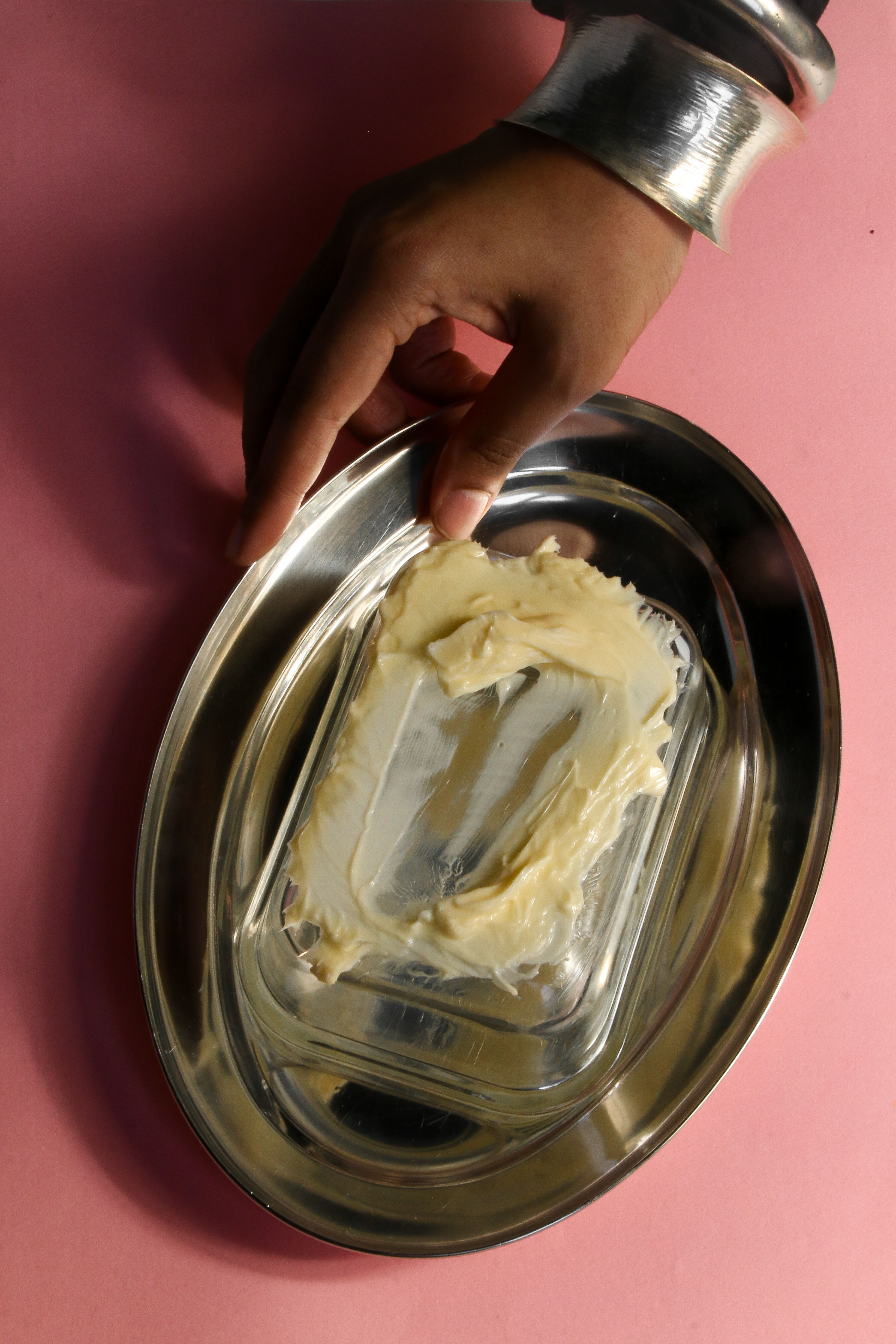 A hand holds a metallic dish with butter on a pink background, showcasing elegance.