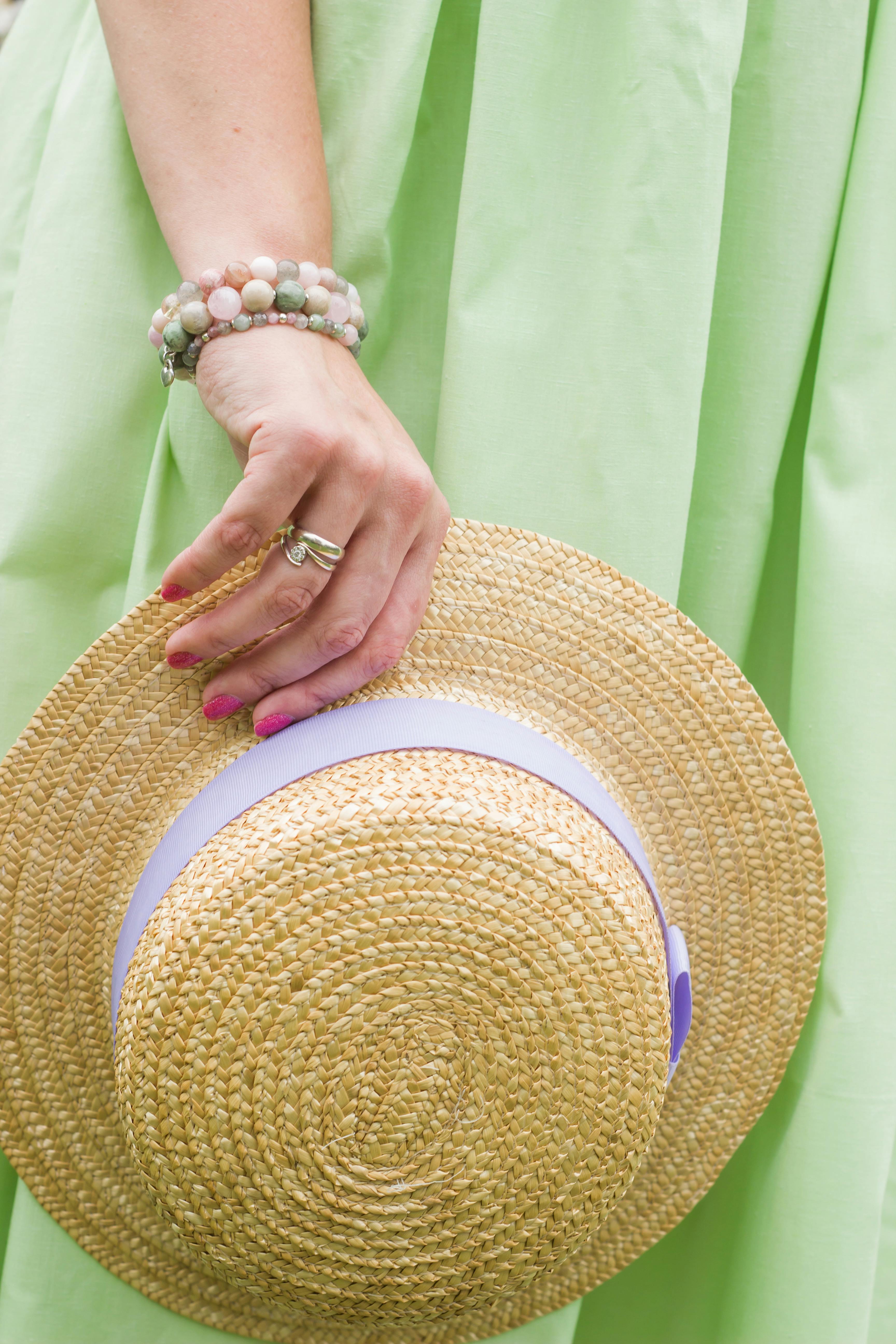 A close-up of an elegant woman holding a straw hat, wearing a green dress and bracelets, perfect for summer fashion themes.