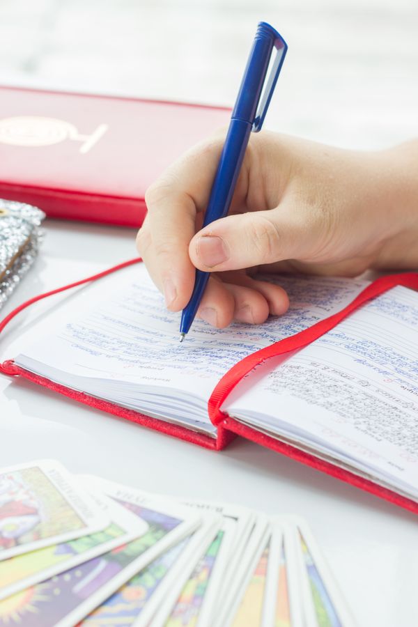 Free Person writing in a red notebook with tarot cards and pen in view. Stock Photo