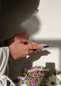 Elegant hands with rings holding smartphone in sunlight with floral arrangement.