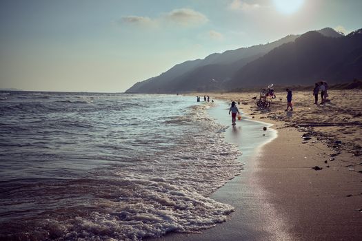 Peaceful beach scene with mountains, people, and gentle waves under a sunny sky.