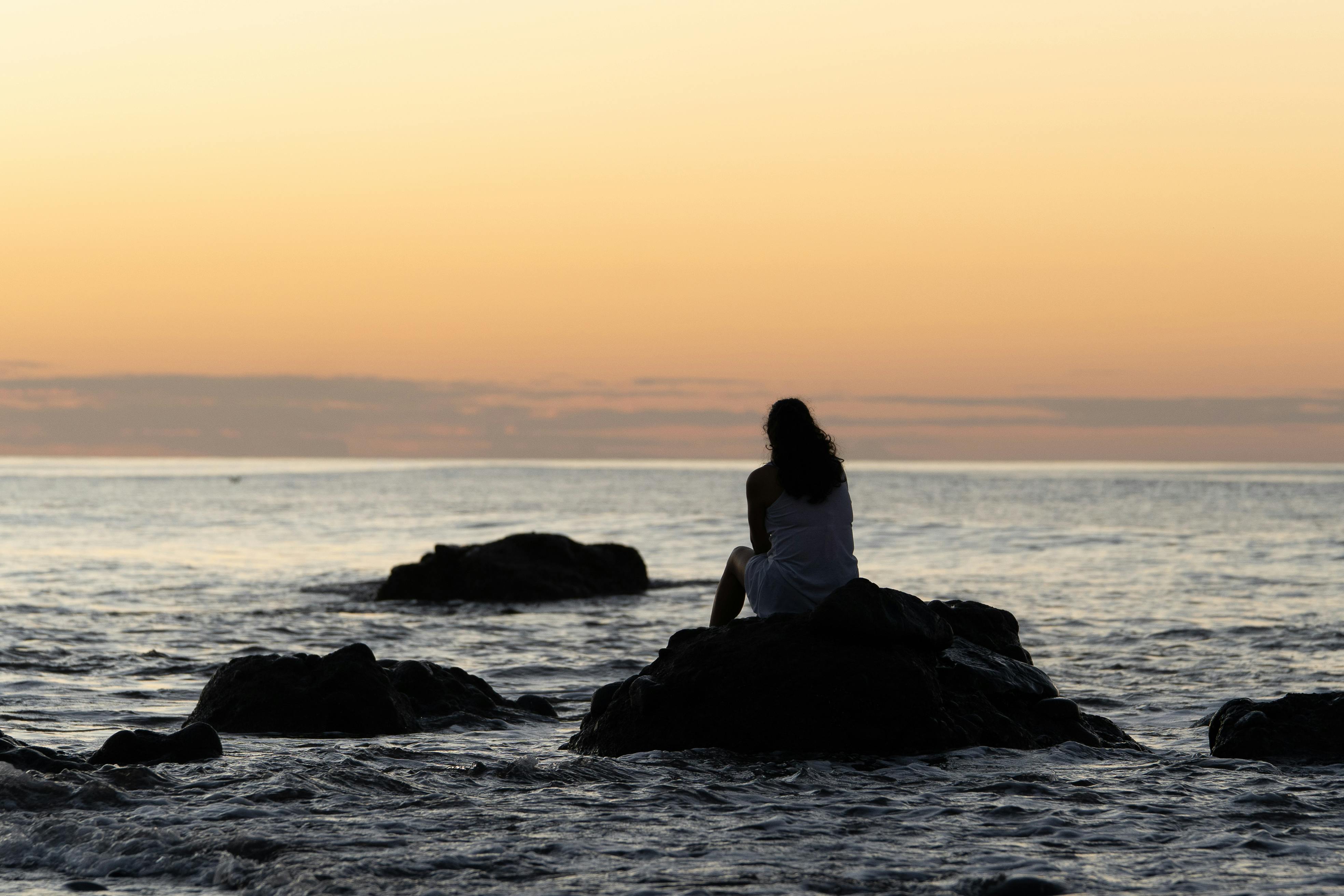 A woman sits on rocks at sunset, contemplating the serene ocean view.