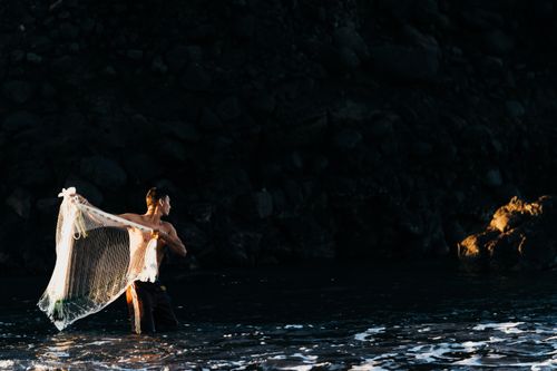 Free A lone fisherman casting a net in twilight by the rocky shore, capturing serene coastal life. Stock Photo