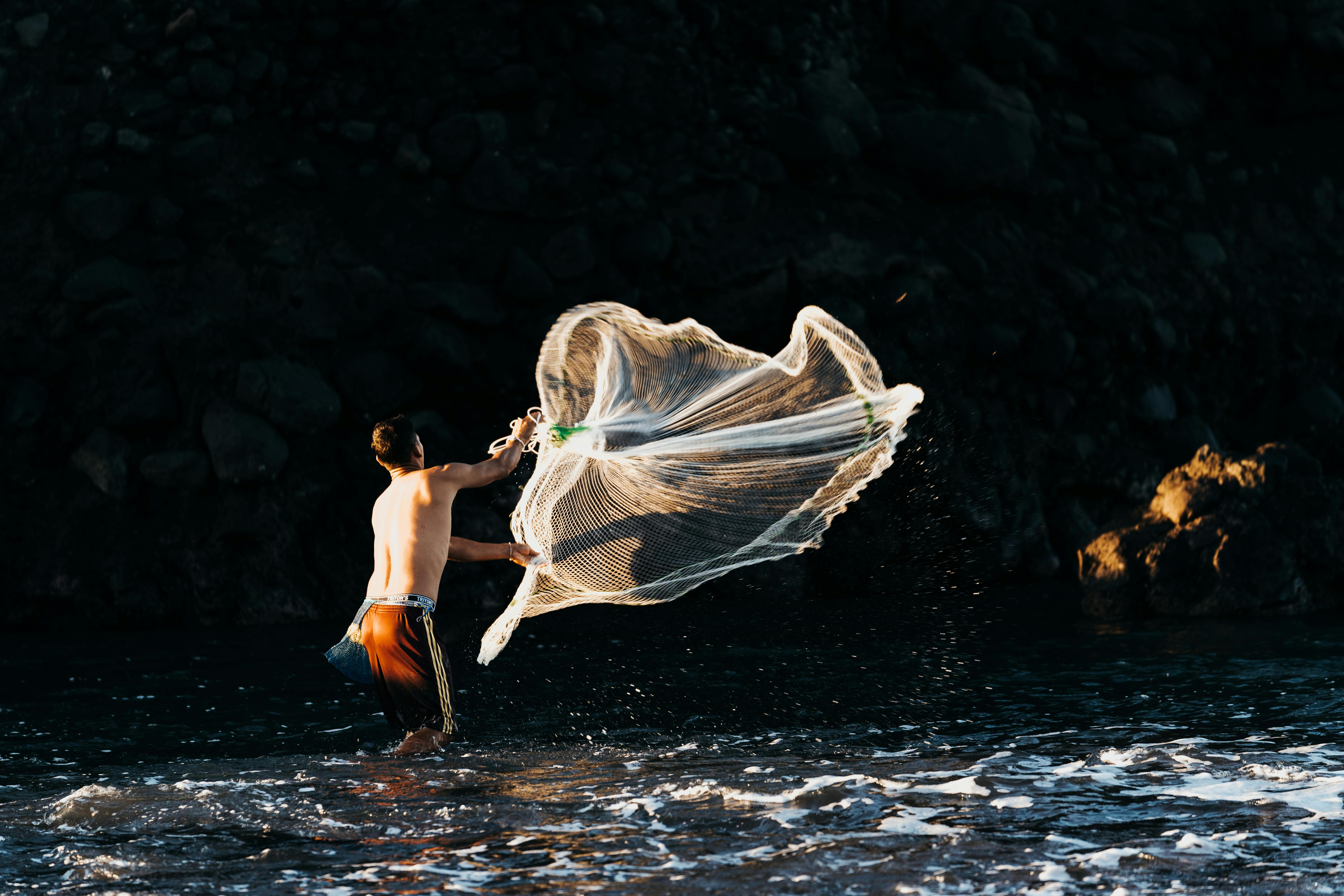 A man throws a fishing net into the ocean at dusk, set against a rocky backdrop.