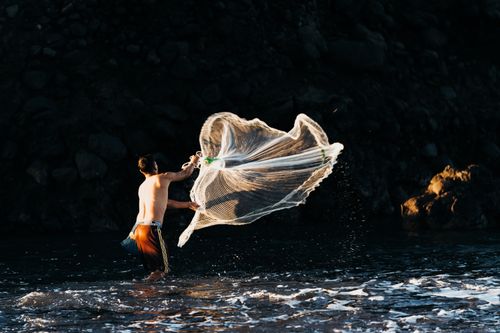 Free A man throws a fishing net into the ocean at dusk, set against a rocky backdrop. Stock Photo