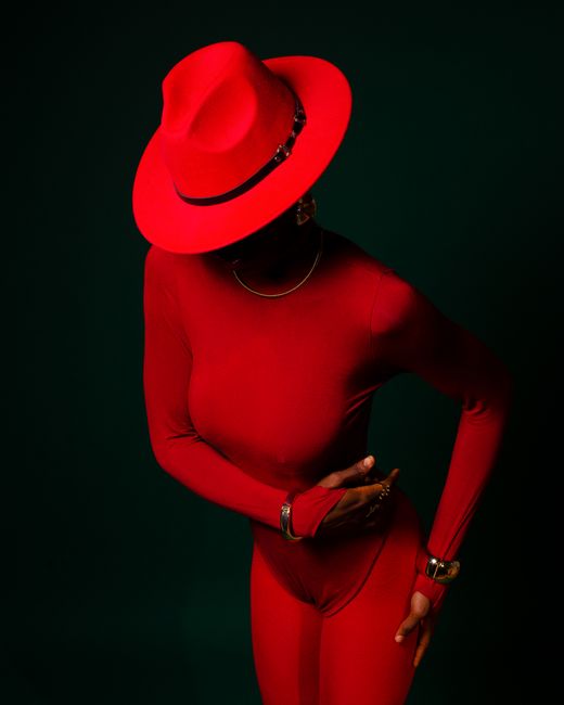 Elegant woman poses in vibrant red outfit and hat against dark background.
