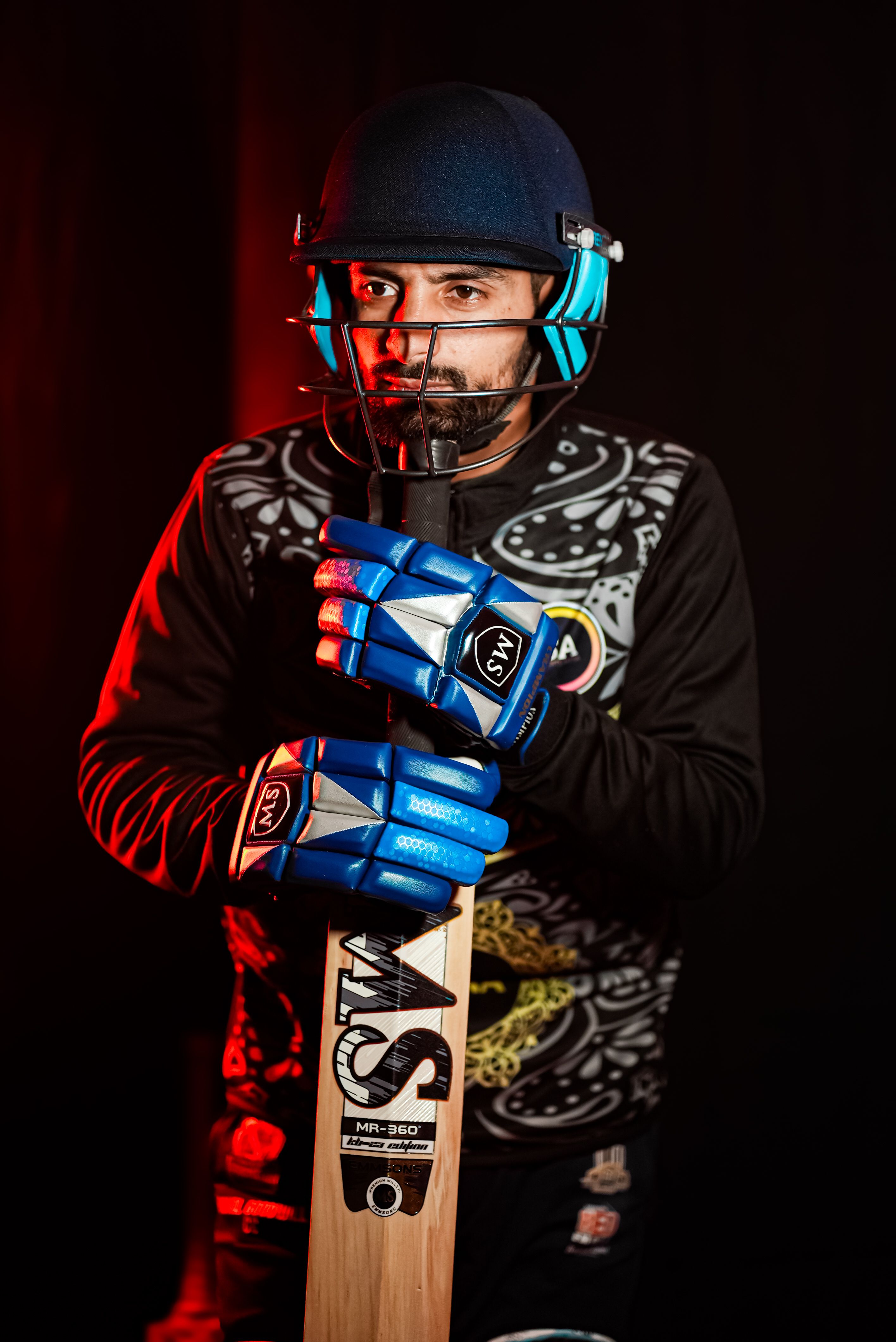 A cricketer wearing protective gear holding a cricket bat, ready for a match under dramatic lighting.