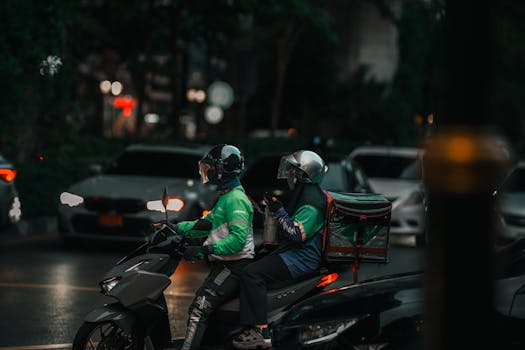 Two delivery riders on a motorbike navigating through city streets at night.