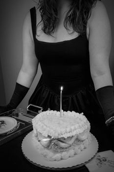 Black and white image of a person in formal attire standing behind a decorated cake with a single lit candle.