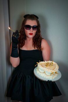 Stylish woman in a black dress holding a birthday cake with number 32 candles indoors.
