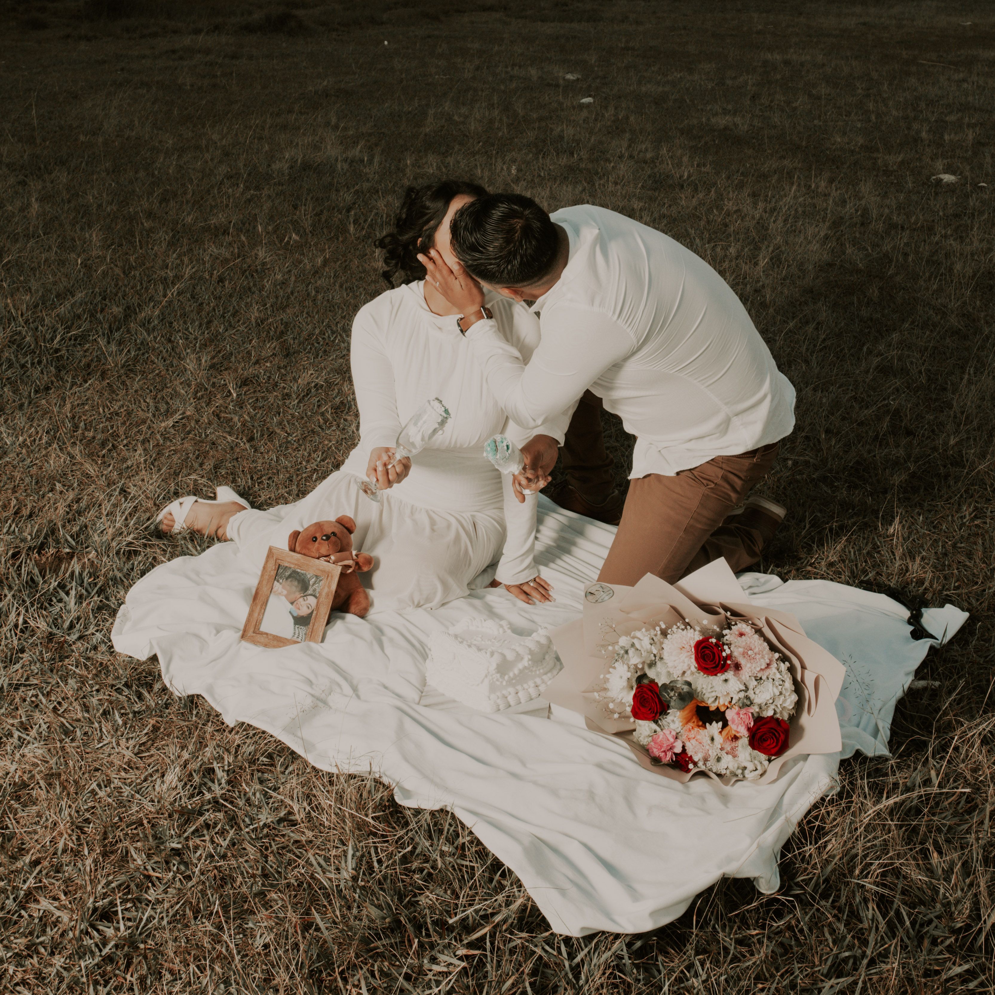 Couple in love sharing tender moment during outdoor picnic with flowers and teddy bear.