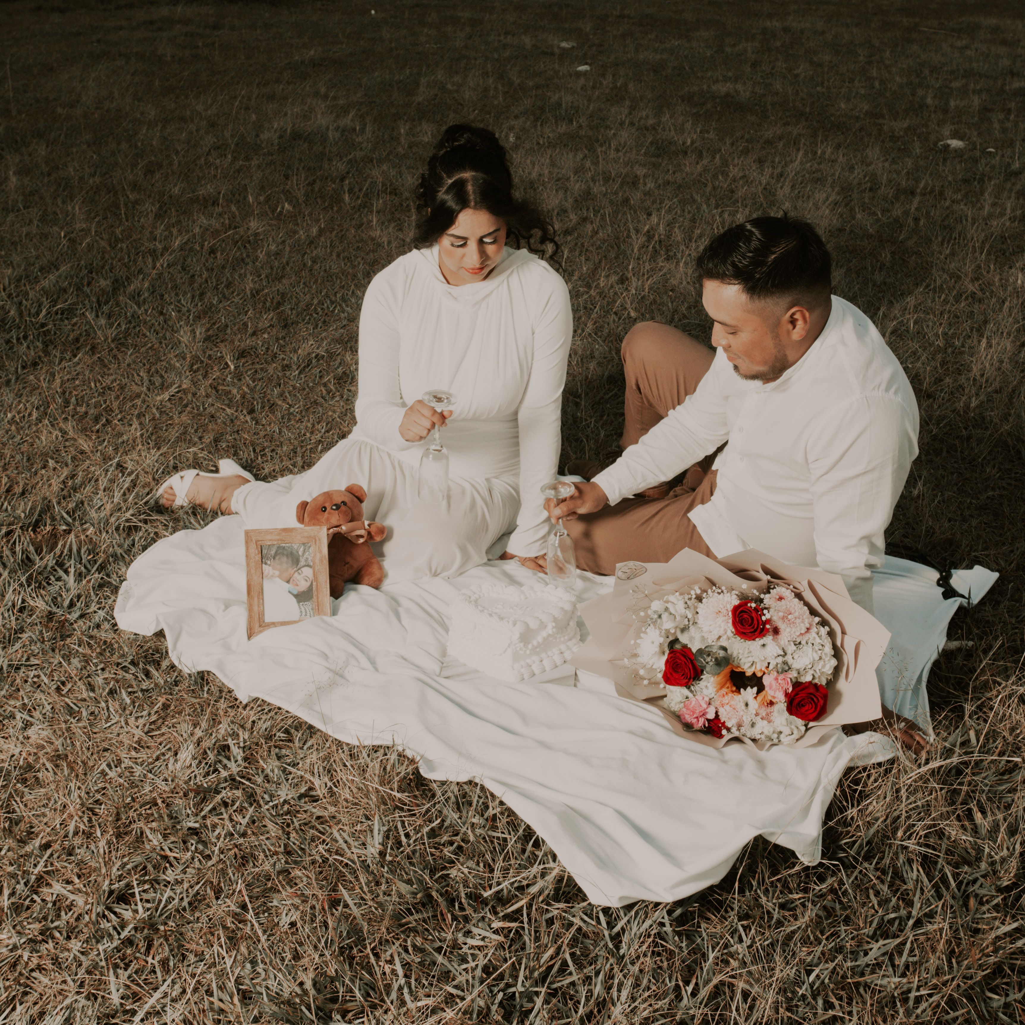 A couple enjoys a romantic picnic on a grassy field, complete with flowers and sentimental items.