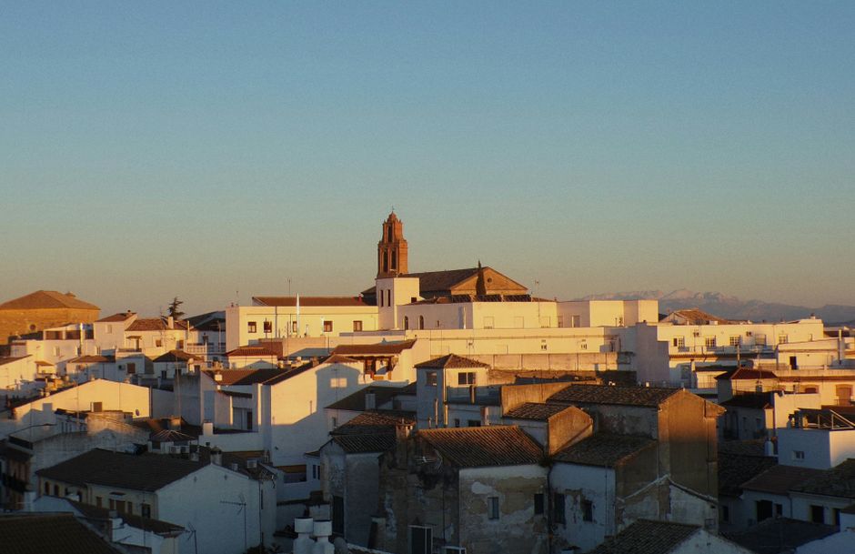 Una vista pintoresca de un pueblo español con edificios blancos al atardecer, destacando una torre de iglesia y montañas distantes.