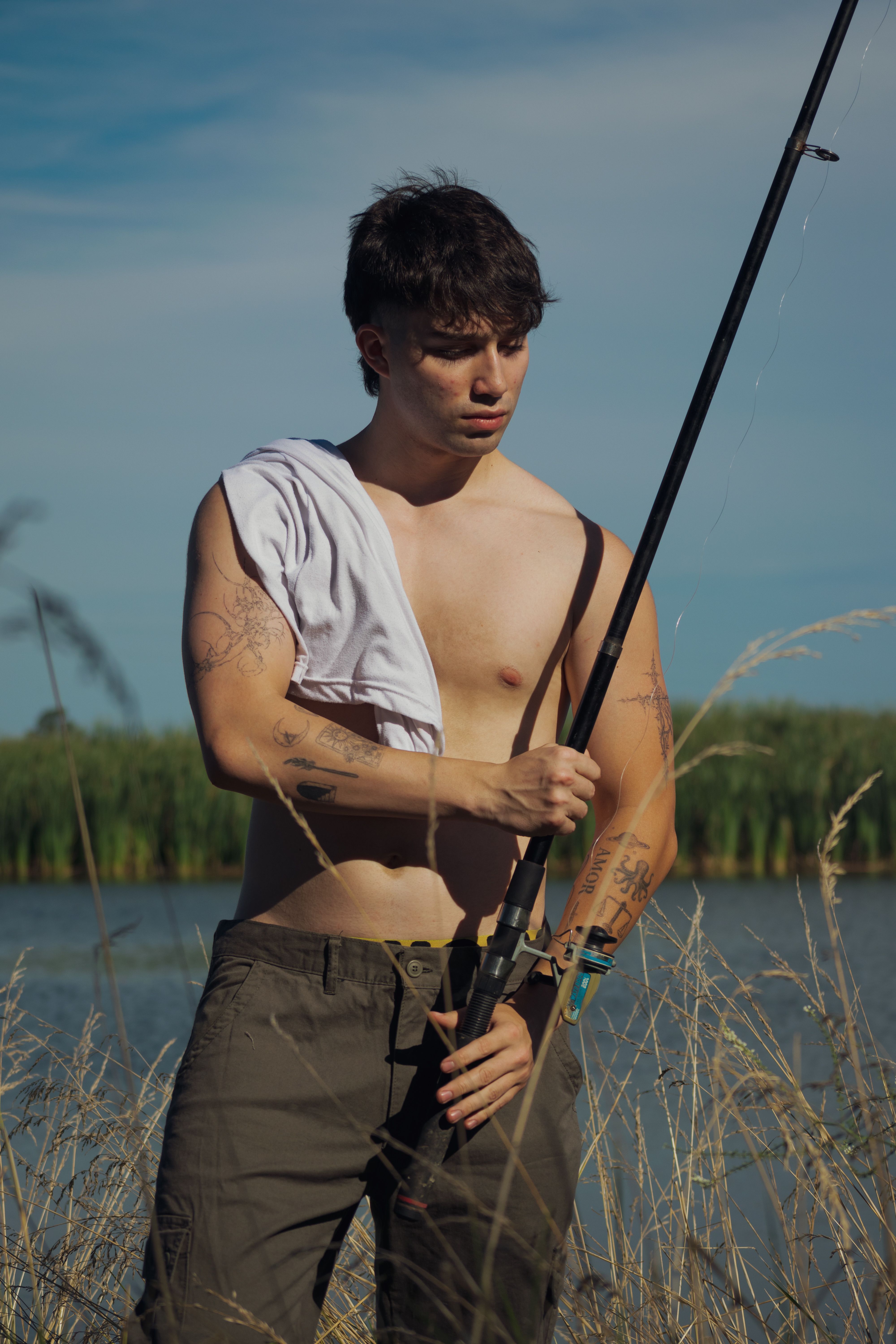 Shirtless young man with tattoos fishing at a serene riverside during a sunny summer day.