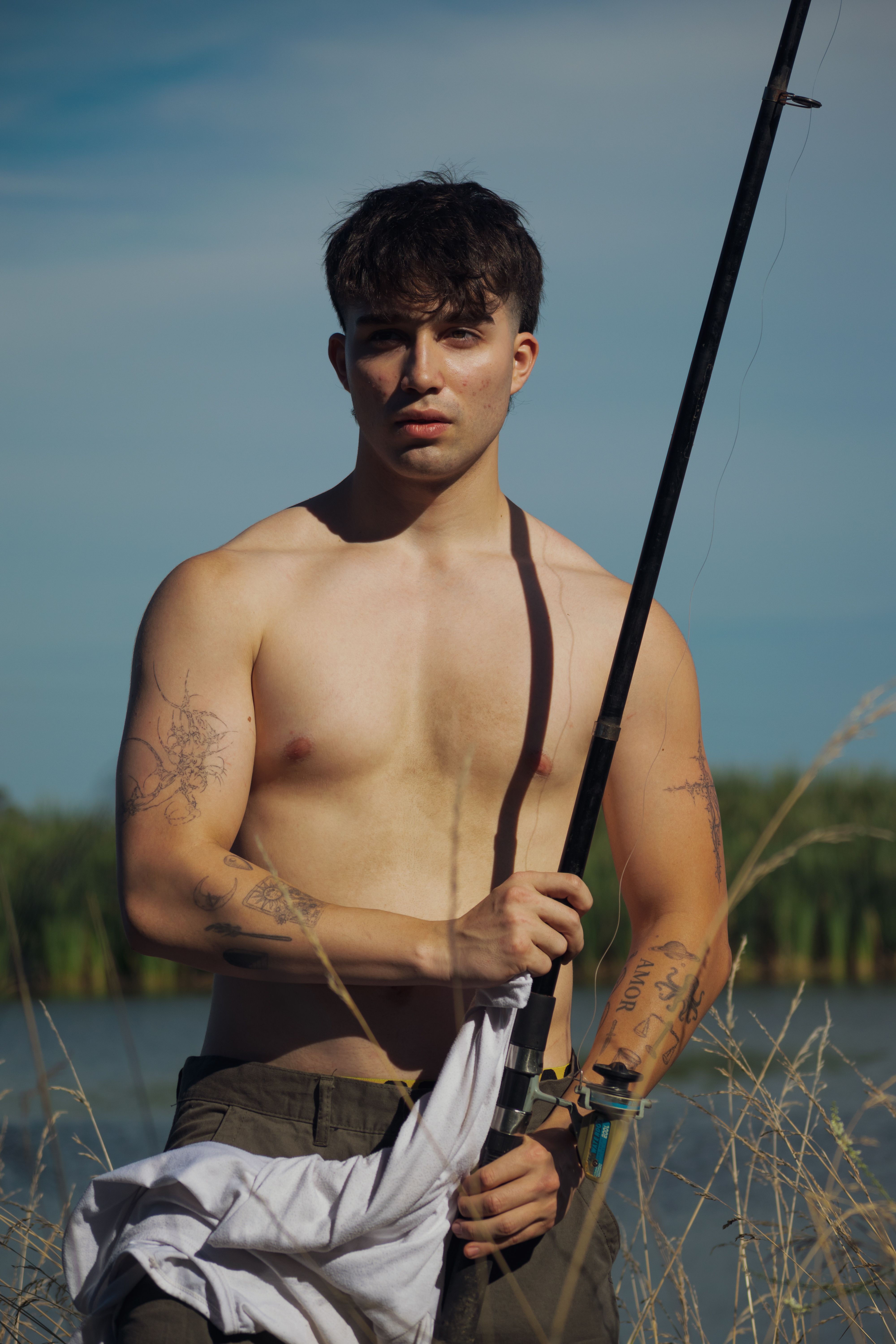 A young shirtless man with tattoos fishes by the river, showcasing strength and serenity.