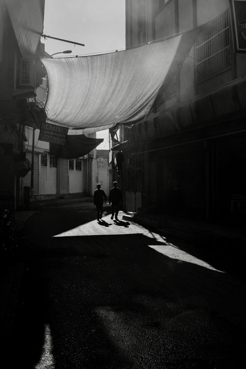 Free Two people walk under hanging fabric in a shadowed urban alley. Black and white photography captures a dramatic scene. Stock Photo