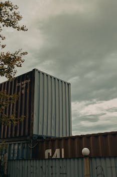 Photo of stacked shipping containers under a cloudy sky in an industrial setting.