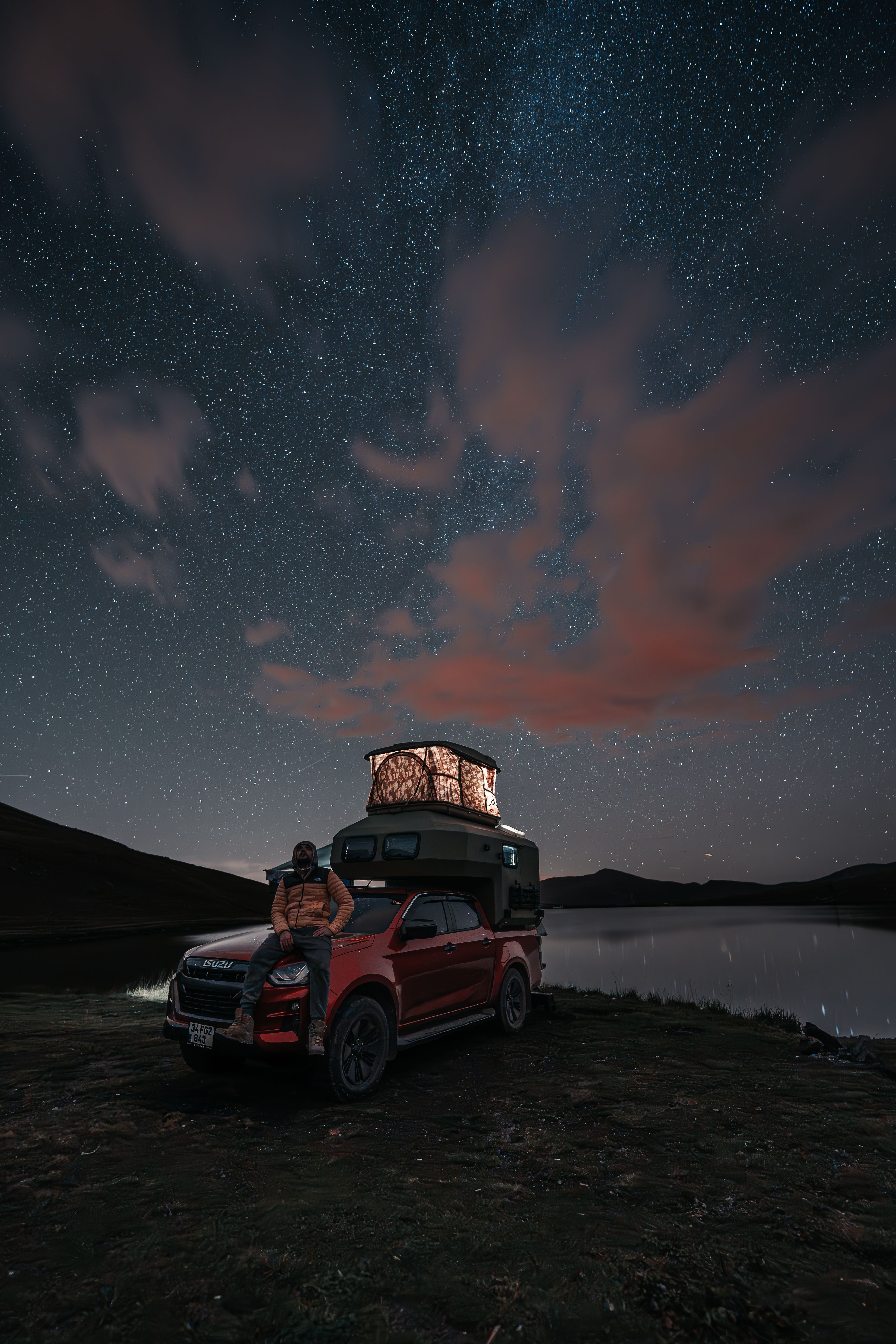 A camper enjoys a starlit sky by a lake with a roof tent on a truck during a nighttime adventure.
