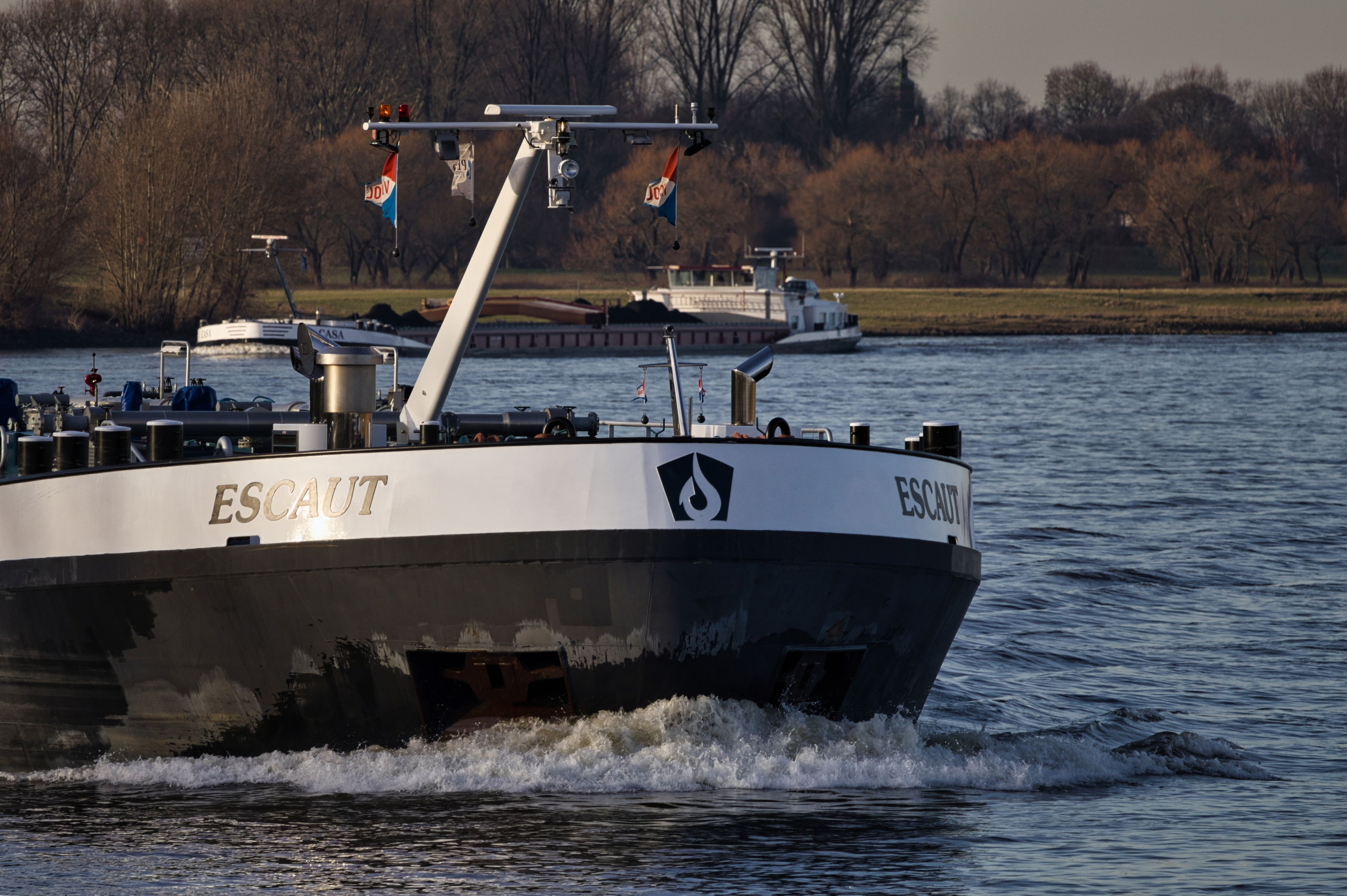 A cargo ship named Escaut navigates a serene river landscape under a clear sky.