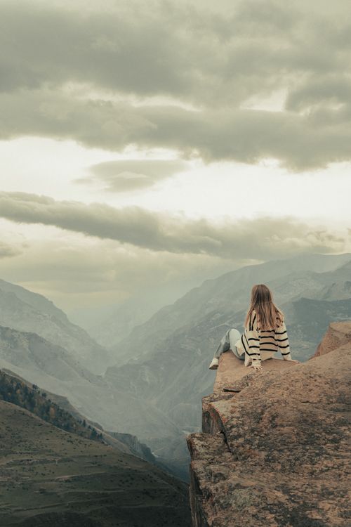 Free Woman sitting on mountain cliff overlooking cloudy valley landscape, serene and contemplative. Stock Photo