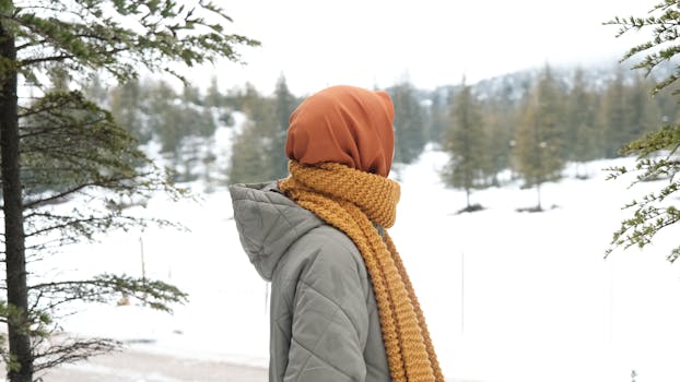 Woman in warm scarf looking at snowy landscape, pine trees around.